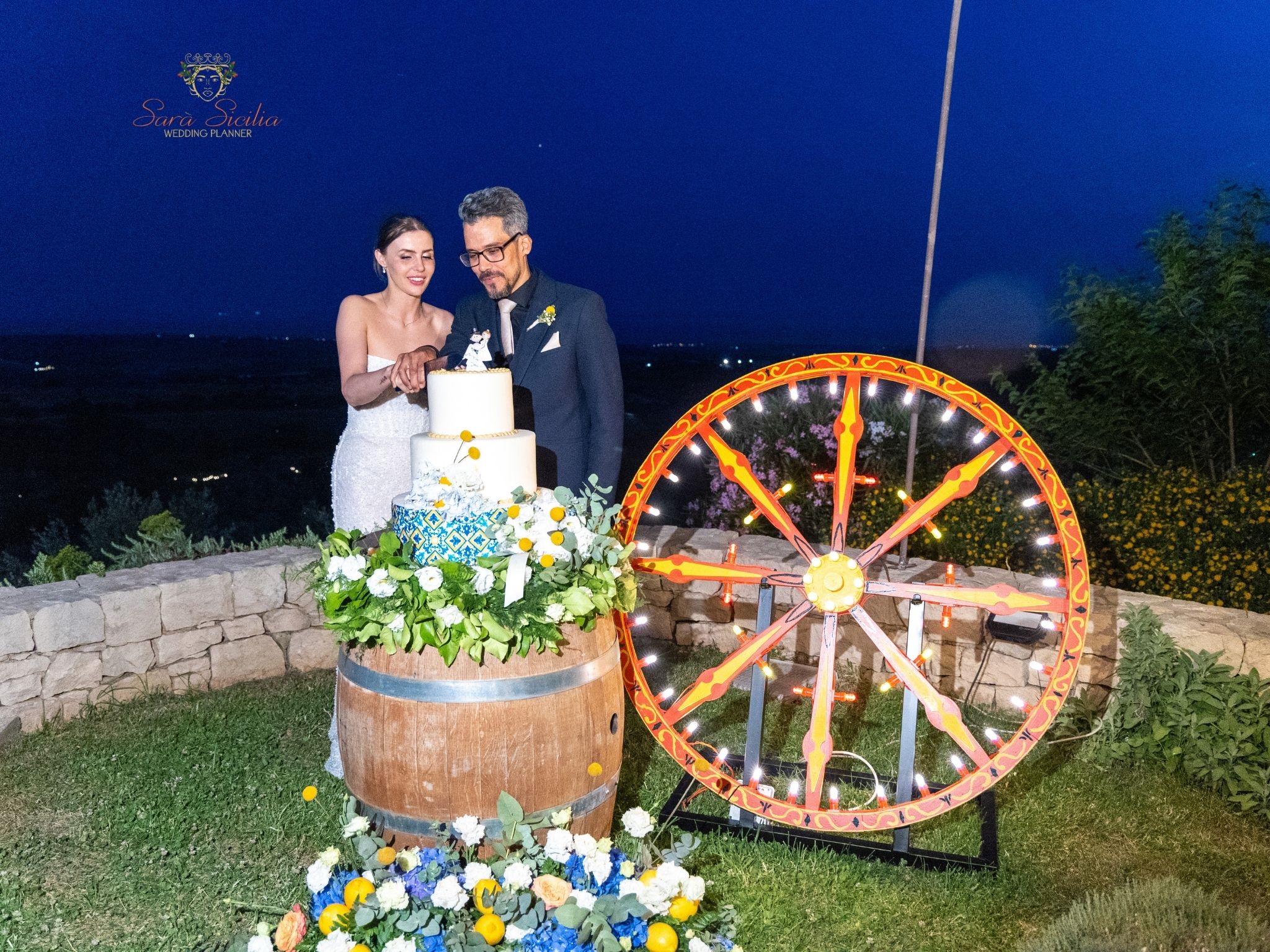 Matrimonio elegante Sicilia – Sposi al taglio torta illuminato da luci romantiche con ruota carretto siciliano
